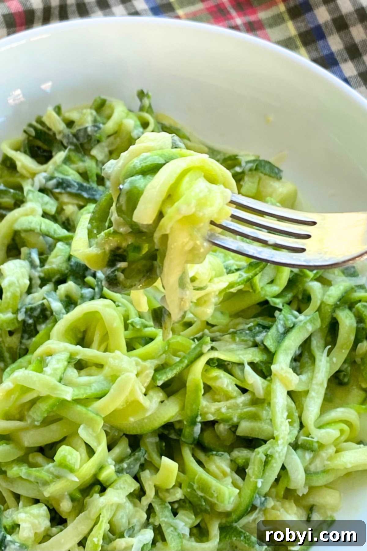A fork lifting a perfect bite of creamy zucchini Alfredo from a white bowl, showcasing the rich sauce coating the noodles.