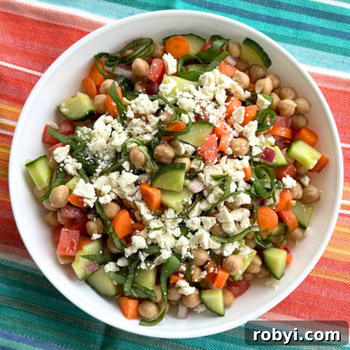 Chickpea feta cucumber salad with red onions, carrots, and tomatoes in a white bowl on a turquoise and orange cloth.