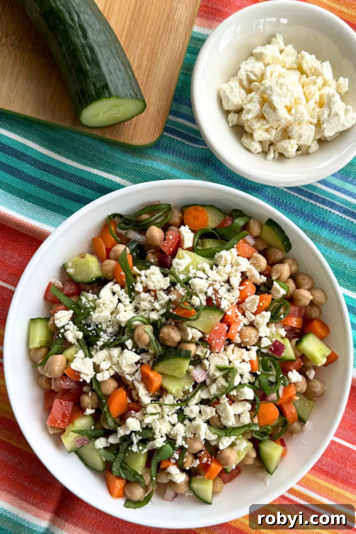 Chickpea Salad in a shallow white bowl with a bowl of feta cheese and an English cucumber behind it.