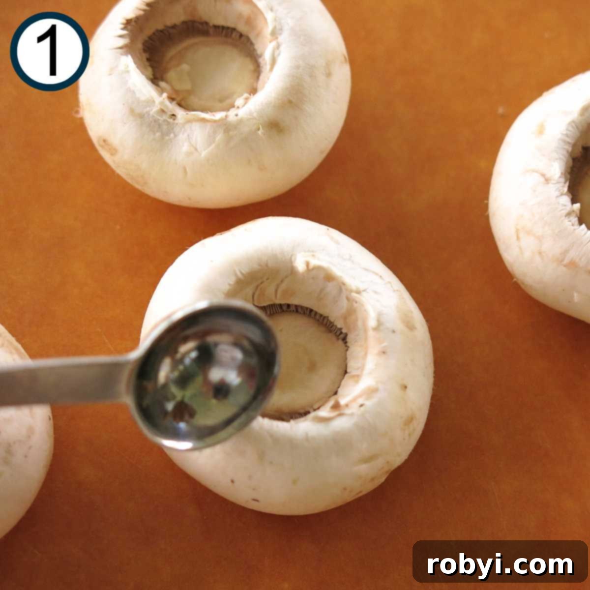 Close-up of a button mushroom being prepped, with its stem removed and the membrane being scooped out using a small measuring spoon.