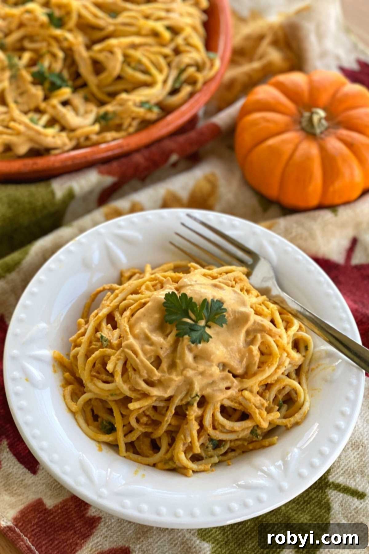 Velvety Pumpkin Pasta Sauce 7 A bowl of spaghetti topped with creamy pumpkin sauce, with whole pumpkins and a decorative platter in the background.