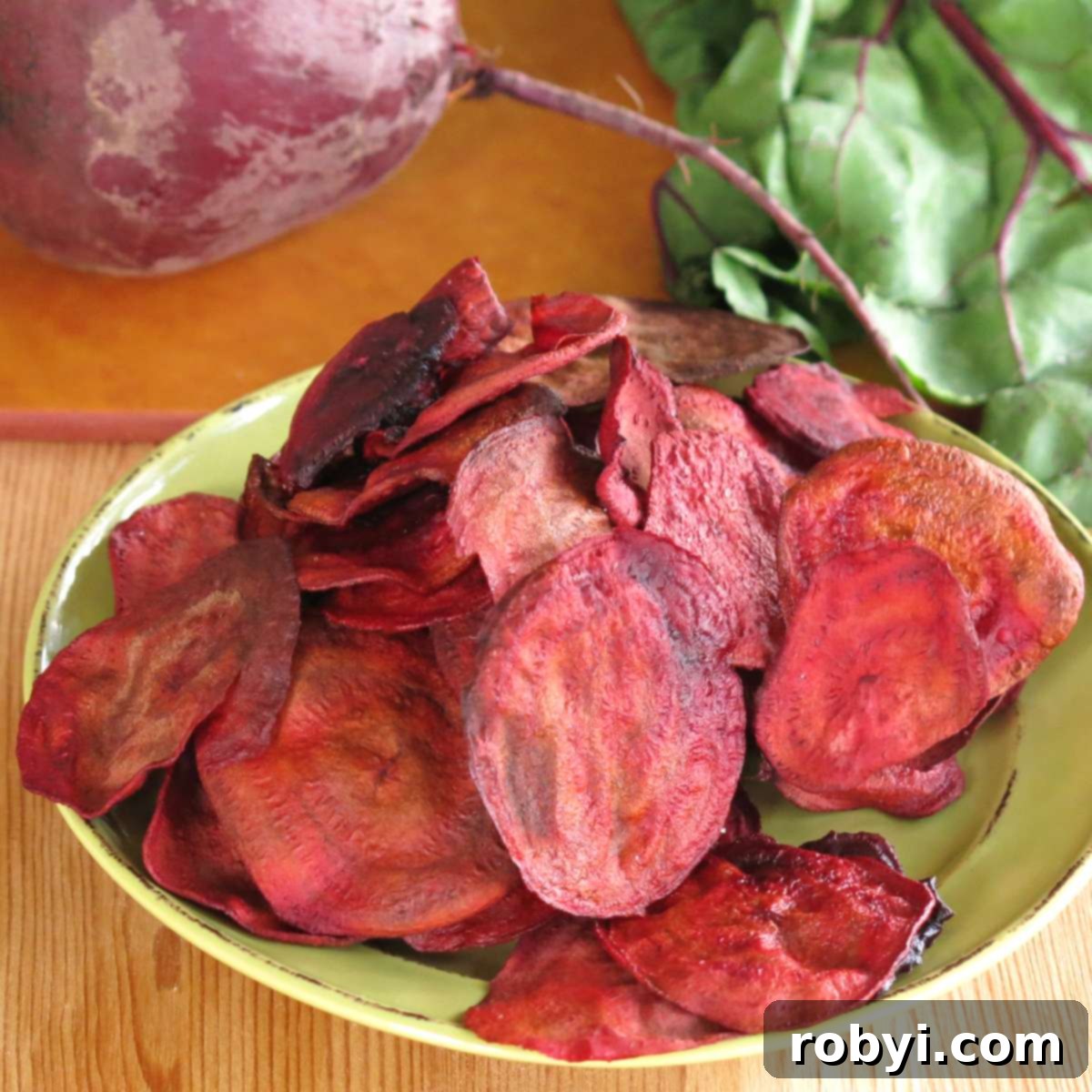Crispy baked beet chips piled high on a vibrant green plate, with a fresh, raw beet and its stem resting behind them, showcasing the natural beauty of the ingredient.