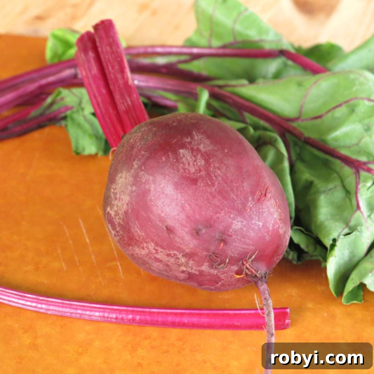 A vibrant red beet rests on a wooden cutting board, showcasing its rich color, with its roots, stems, and leaves still attached, ready for preparation.