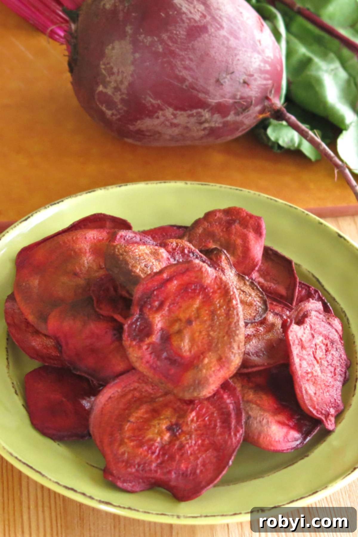 A vibrant pile of homemade baked beet chips sits on a white plate, with a whole raw beet nestled behind them on a rustic cutting board, highlighting the freshness of the ingredients.