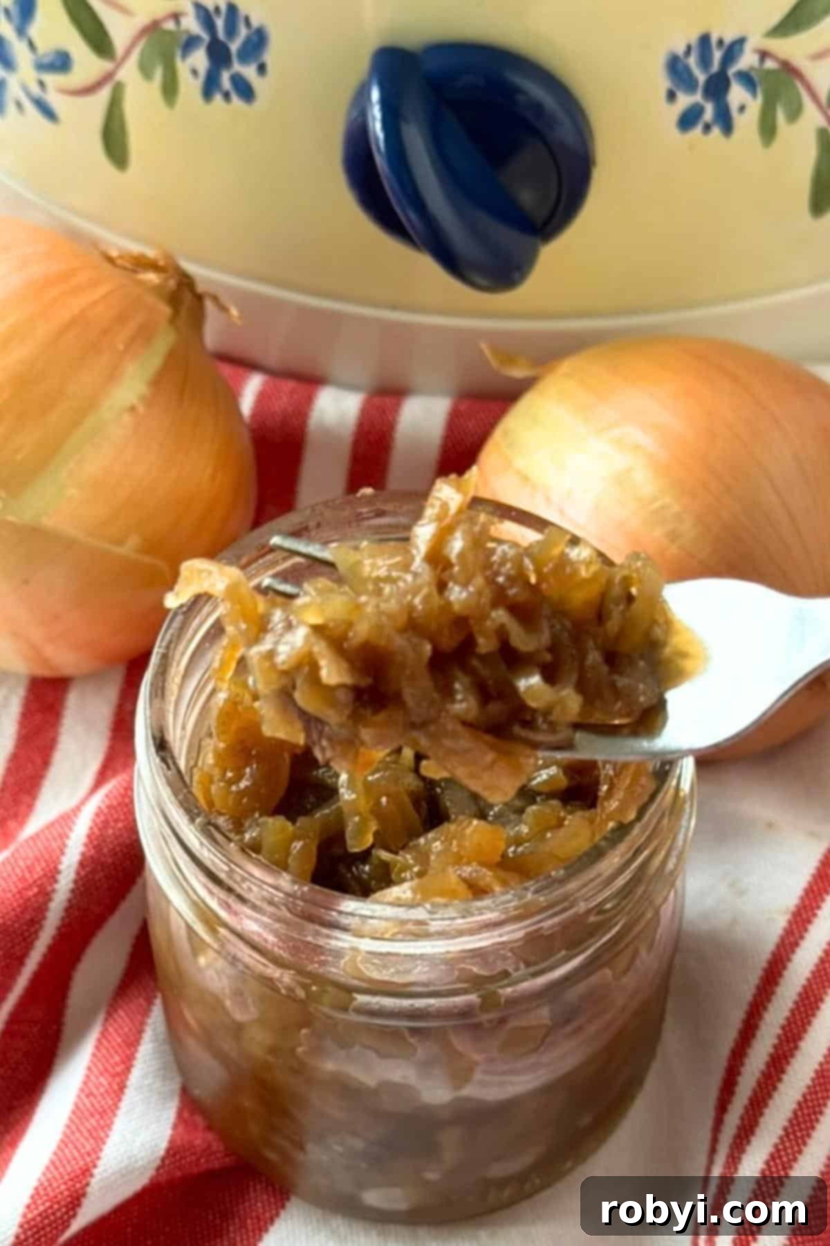 A fork lifting caramelized onions out of a glass jar, with two yellow onions and a slow cooker in the soft background.