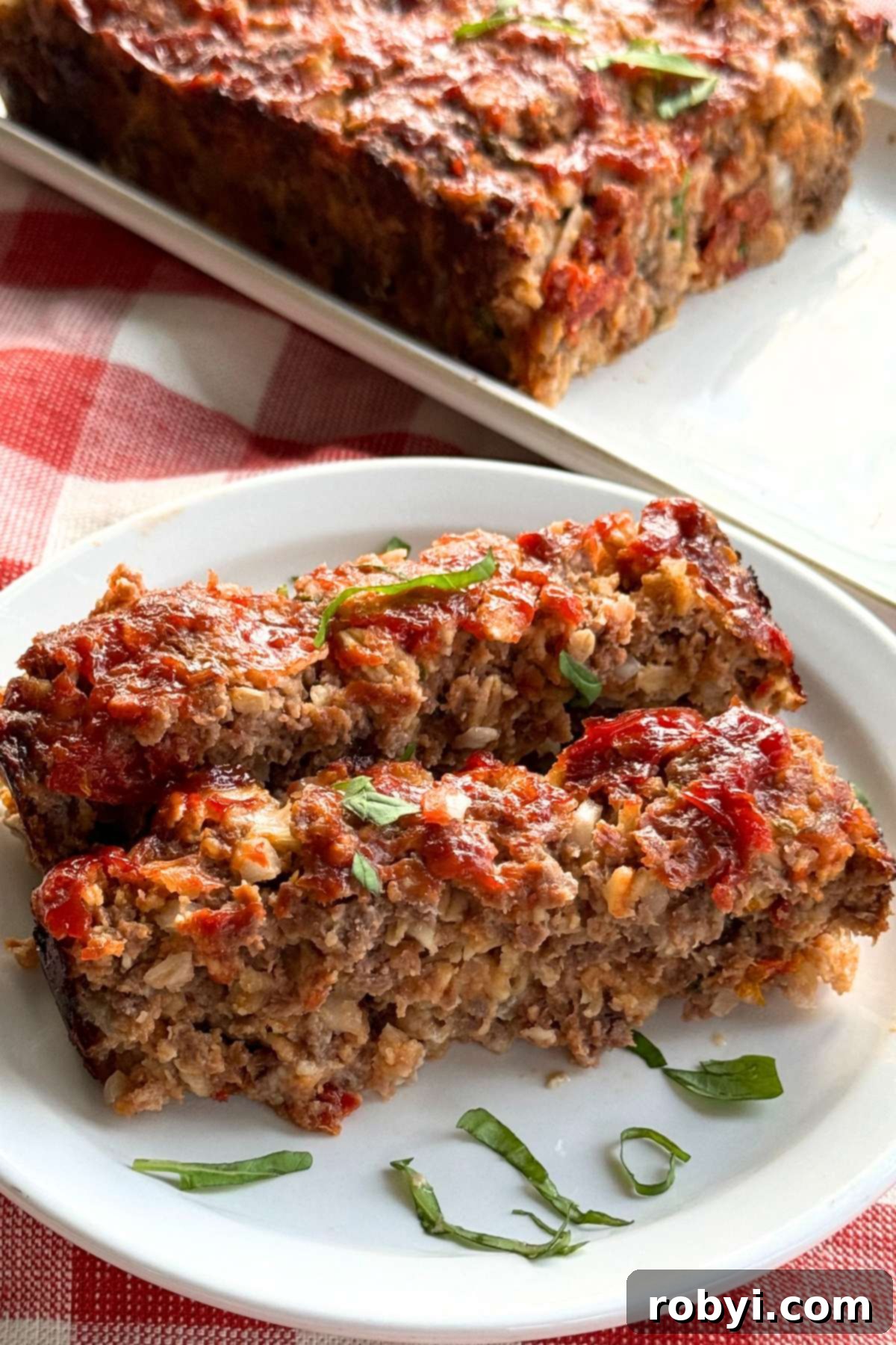 Two slices of sundried tomato meatloaf made with oatmeal on a white plate with chopped basil, with the rest of the loaf behind it. The meatloaf is rich in color with visible pieces of sun-dried tomatoes and herbs.