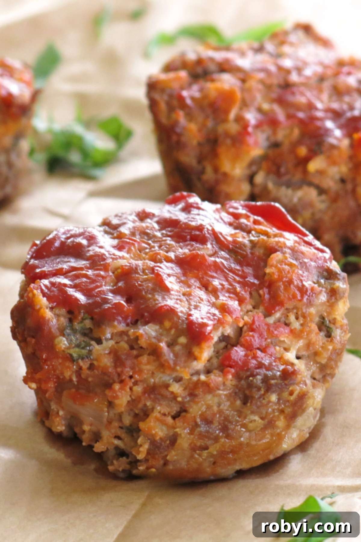 Two gourmet meatloaf muffins made with oatmeal, sun-dried tomatoes, and cheese on brown paper scattered with fresh basil, showcasing their individual portions and delicious crust.